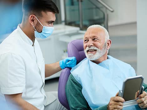 Dentist in mask and gloves shows tablet results to smiling senior patient in dental office.