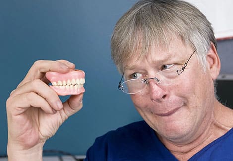 Dentist wearing glasses holding dental model against blue background in clinical setting.