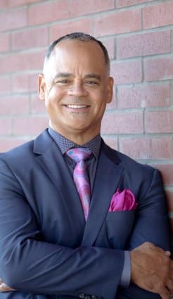 Professional headshot of smiling man in navy blazer and purple shirt against brick wall background.
