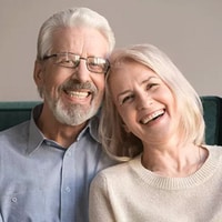 Smiling older couple wearing glasses in casual clothing against neutral background.