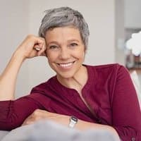 Smiling woman with short gray hair in burgundy top, hand near face, indoor setting.
