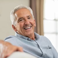 Smiling older man in light blue shirt, indoors with window background, dental care setting.