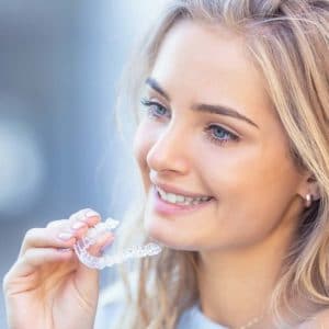 Smiling woman holding clear aligner near mouth in bright dental setting.