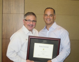 Two men hold framed certificate in professional dental office setting.