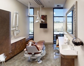 Modern dental operatory with patient chair, overhead light, sink, and large windows overlooking cityscape.
