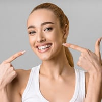 Young woman pointing to her smile against gray background, promoting dental health and care.