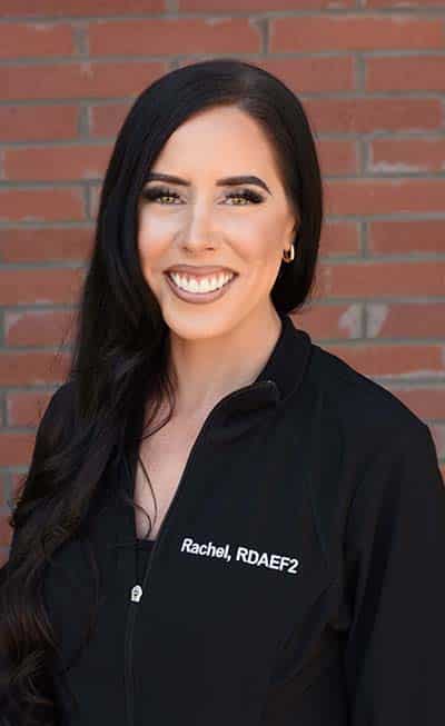 Professional female dentist smiling at camera wearing black scrubs against brick wall background.