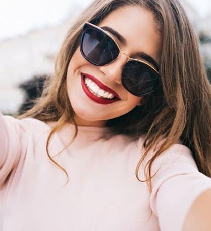 Woman wearing sunglasses and red lipstick smiling outdoors in casual pink top.