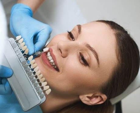 Dentist holding tooth shade guide against patient's teeth during smile assessment consultation.