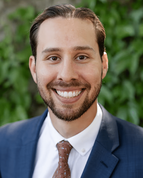 Professional male dentist smiling at camera wearing blue blazer and tie against green background.