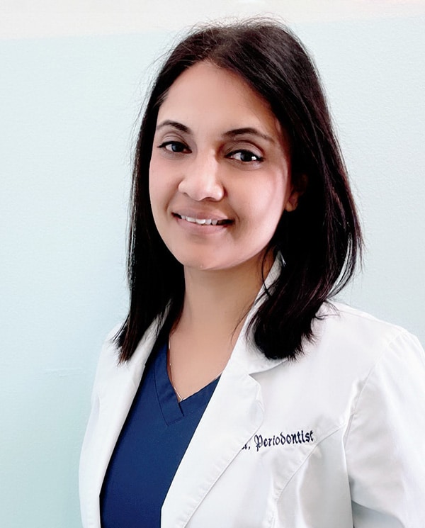 Female dentist in white coat and blue scrubs smiling at camera against white background.